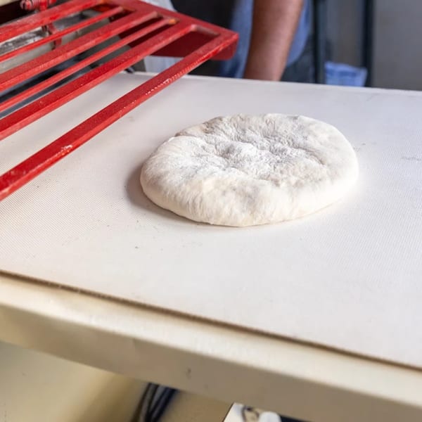 A round, floured Buzzi's Pizza dough sits on a white surface next to a red pizza peel.