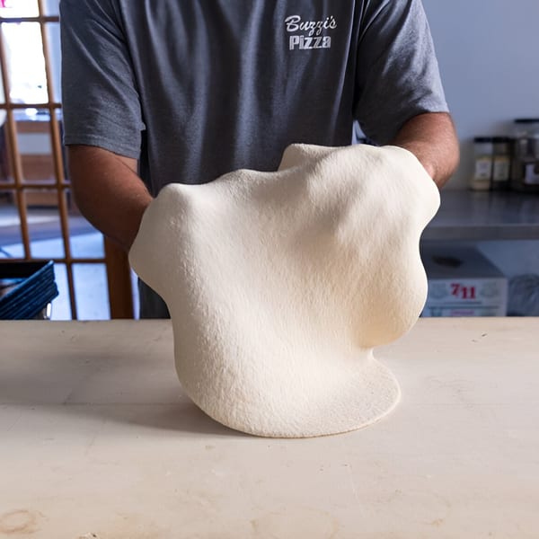 A Buzzi's Pizza chef stretches fresh pizza dough over a light-colored counter.