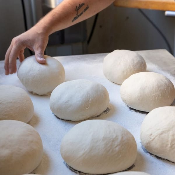 A hand with a tattoo on the forearm gently presses a round, white pizza dough ball on a floured surface.