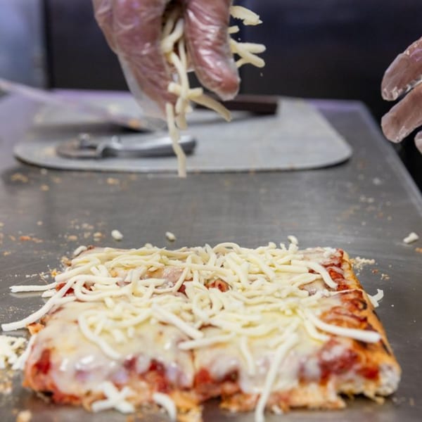 Gloved hands sprinkle shredded mozzarella cheese onto a square slice of Buzzi's Pizza.