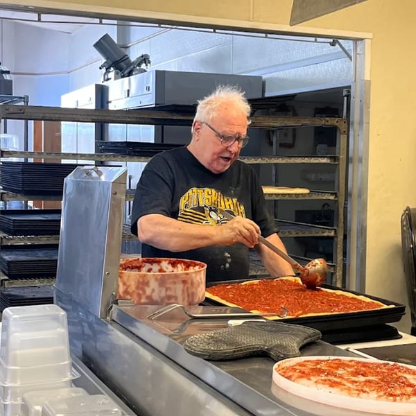 An older man in a Buzzi's Pizza shirt spreads red sauce on a large pizza in a kitchen.