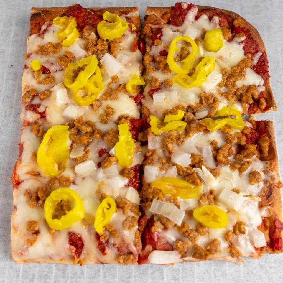 Four square slices of Buzzi's Pizza with sausage, onions, and yellow peppers on a white surface.