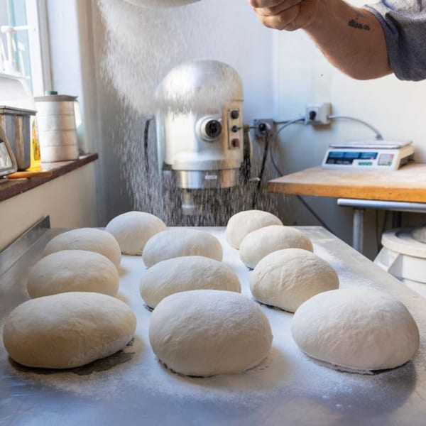 A baker's hands dust flour over rows of Buzzi's Pizza dough balls on a metal table.
